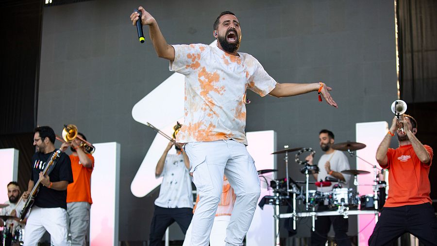 Un cantante con camiseta blanca y pantalones blancos, con los brazos extendidos y un micrófono, actúa en un escenario frente a una banda. Se observan músicos tocando instrumentos como bajo, trombón, trompeta y batería.