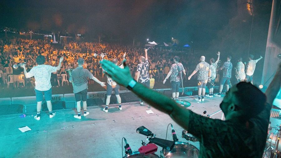 Un concierto al aire libre de la banda La Fúmiga, con los músicos de espaldas al público y una multitud entusiasta. Se distinguen instrumentos musicales y luces de escenario.