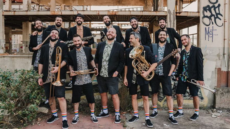 Un grupo de músicos posa en un entorno urbano con grafitis, vistiendo chaquetas negras, camisas estampadas y pantalones cortos. Llevan calcetines de arcoíris y sostienen instrumentos como trompetas y trombones.