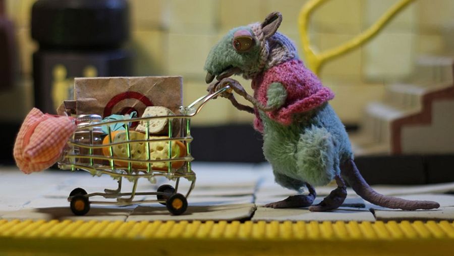 Una rata de color verde azulado con ropa rosa y gris empuja un carrito de compras lleno de objetos, como una bolsa con un logotipo, un tarro y comida. El fondo muestra una pared de azulejos y una escalera.