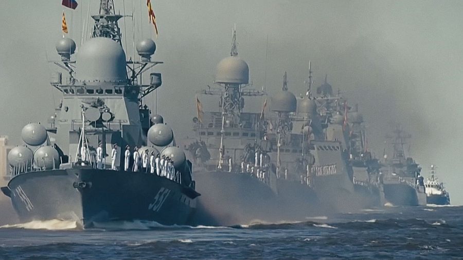 Una flota de barcos grises navega por un mar agitado. Se observan personas vestidas con uniformes blancos a bordo de los barcos, y banderas ondean en los mástiles. El número 