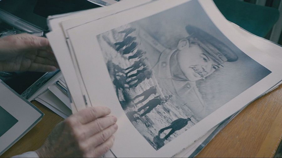 Unas manos sostienen fotografías antiguas, incluyendo un retrato de un hombre con bigote y uniforme militar, y una imagen de personas en un paisaje nevado, evocando una atmósfera sombría.