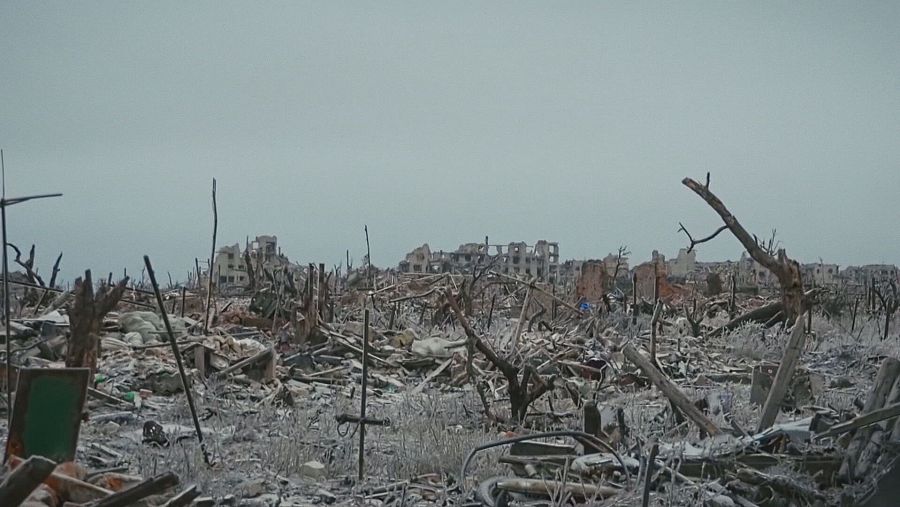 Una escena de destrucción muestra edificios en ruinas y escombros, con restos dispersos por el suelo y vegetación dañada, dominada por tonos grises que evocan una atmósfera de desolación.
