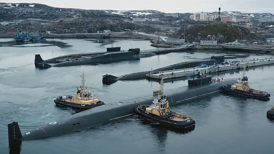 En un fiordo, un submarino de gran tamaño es remolcado por dos embarcaciones. Otros submarinos se encuentran en el agua, y al fondo se distingue una ciudad costera.