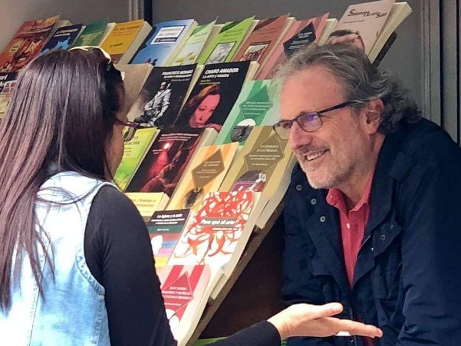 Carlos de la Cruz, con gafas y camisa roja, interactúa con una mujer en la Feria del Libro, con una estantería llena de libros como telón de fondo. La mujer lleva una chaqueta vaquera sin mangas.