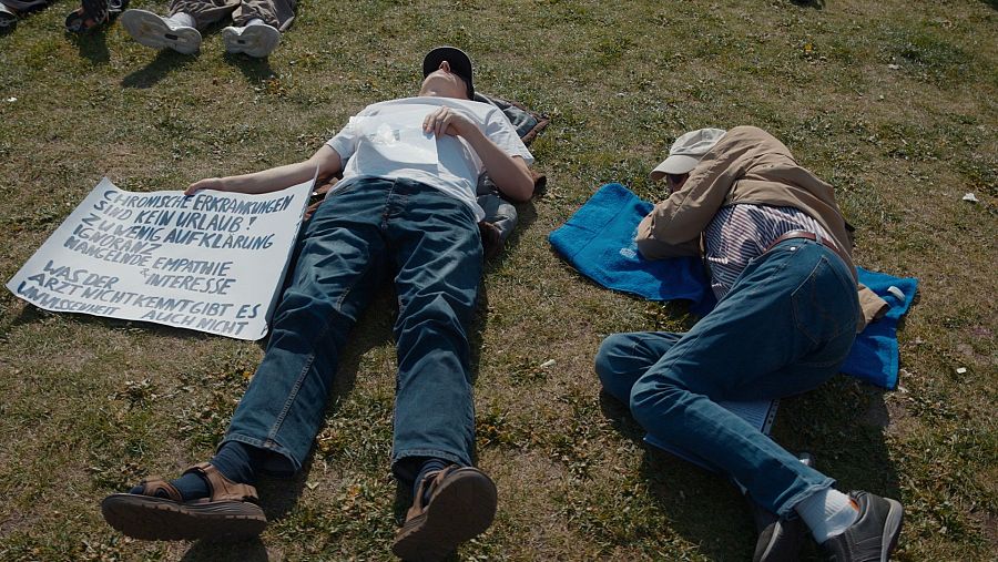 Dos personas yacen en el césped, una con una pancarta en alemán y otra sobre una toalla azul. La pancarta critica la falta de conocimiento y empatía hacia enfermedades crónicas, posiblemente en el contexto de una protesta.