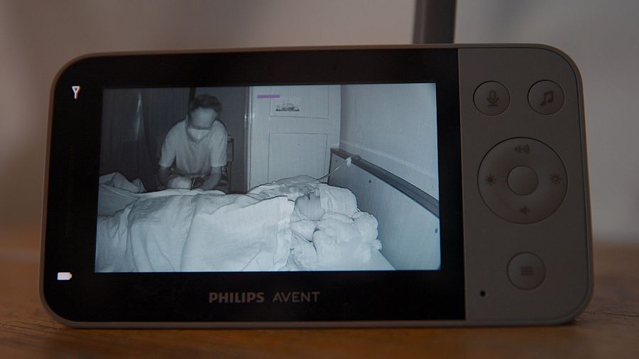 Un monitor de bebé Philips Avent muestra una habitación con una cama y una persona cuidando a otra que está acostada y cubierta con una manta, en una imagen en blanco y negro.