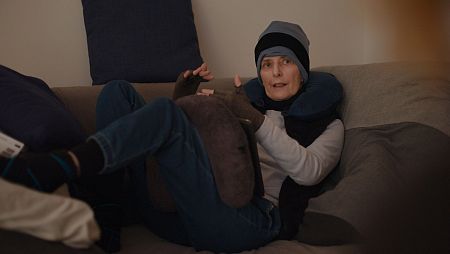 Una mujer con gorro, guantes sin dedos y un cojín de viaje, se encuentra sentada en un sofá con las piernas cruzadas, usando pantalones vaqueros y calcetines. Tiene un cojín sobre las piernas.