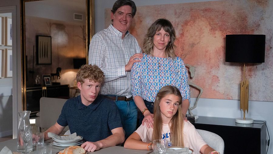 Una familia posa en el salón de su casa. Los padres, vestidos con ropa informal, están de pie detrás de sus hijos, quienes están sentados a la mesa. En la mesa hay platos y vasos.