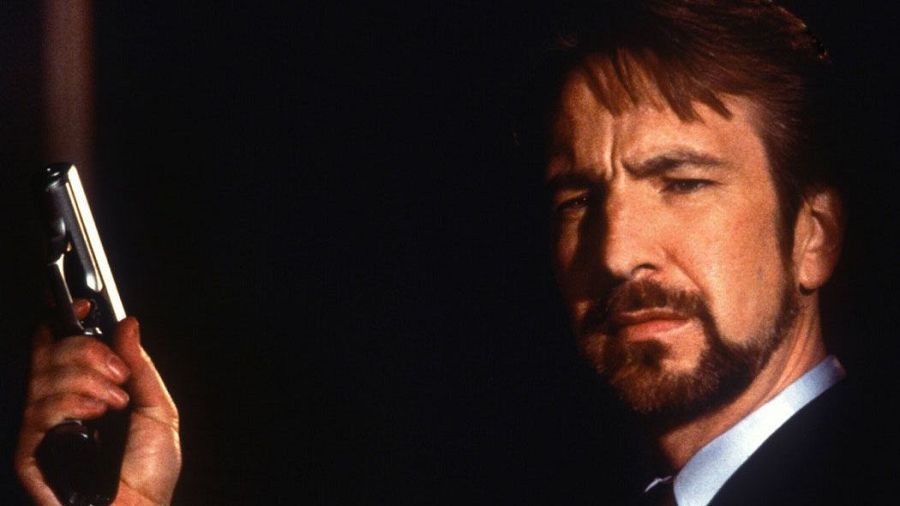 Alan Rickman, con el cabello peinado hacia atrás y barba de candado, sostiene una pistola plateada. Viste un traje oscuro con camisa blanca, con una expresión seria y la mirada fija.