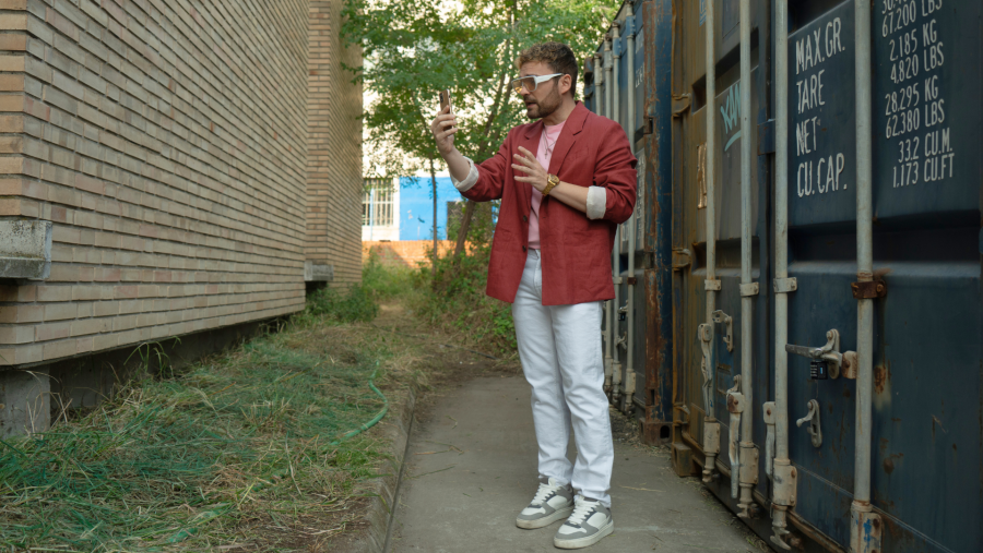 Adam Jezierski, como Fred, se encuentra en un callejón, hablando por videollamada. Viste ropa casual: chaqueta roja, camiseta rosa, pantalones blancos y zapatillas deportivas. A su alrededor hay contenedores de carga y vegetación.