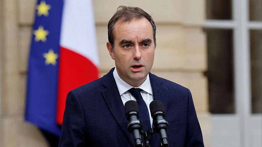 En la imagen, Sébastien Lecornu, con traje y corbata, habla ante dos micrófonos. Se encuentra frente a la bandera de Francia, posiblemente comentando la situación política del país.