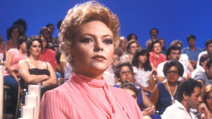 Mayra Gómez Kemp, con un vestido rosa y cabello rubio rizado, posa frente a un público numeroso. Destaca su maquillaje, con énfasis en ojos y labios, en un fondo azul.