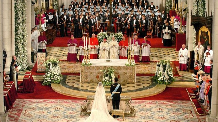 En la iglesia, se observa a los novios, ella con vestido blanco y velo, él con uniforme militar, frente a un altar adornado con flores. Detrás, se distinguen obispos y un coro, con invitados a los lados.