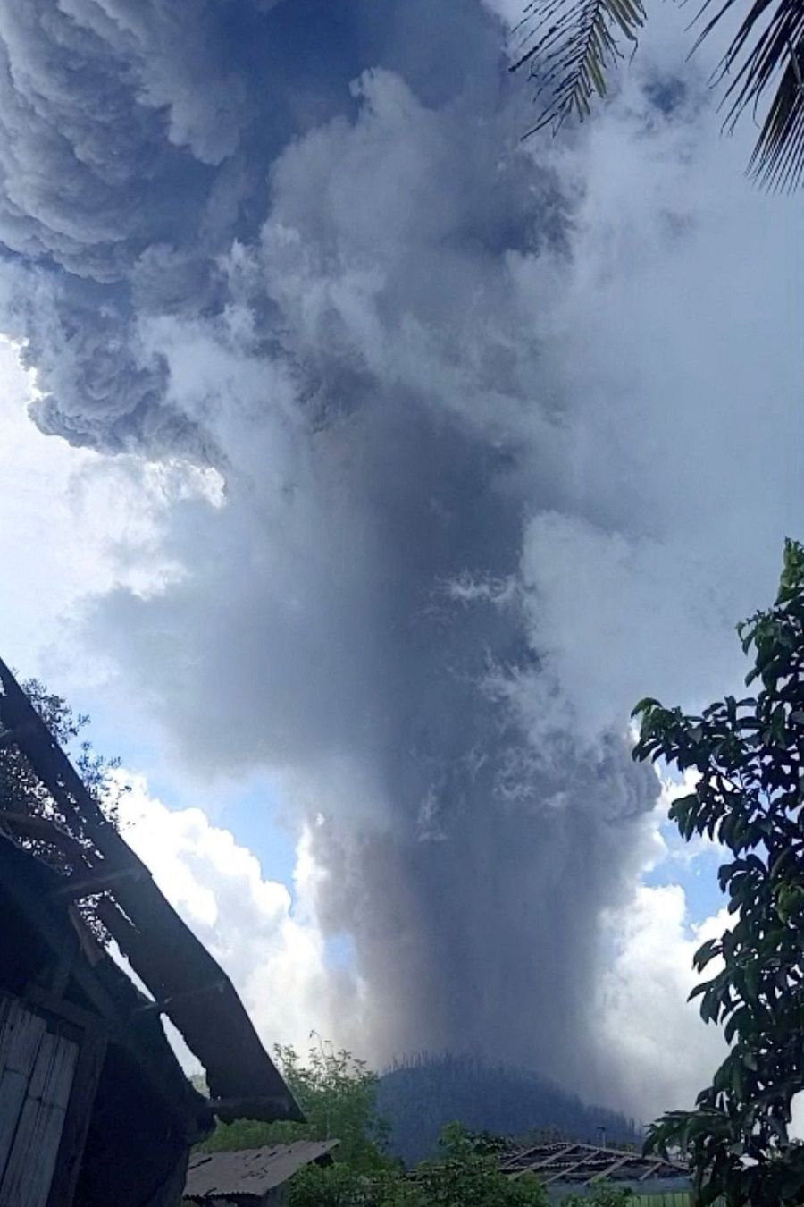 El monte Lewotobi Laki-laki, en Indonesia, entra en erupción, lanzando una densa columna de humo y ceniza al cielo. Se observan estructuras y vegetación en primer plano, con el cielo parcialmente cubierto de nubes.