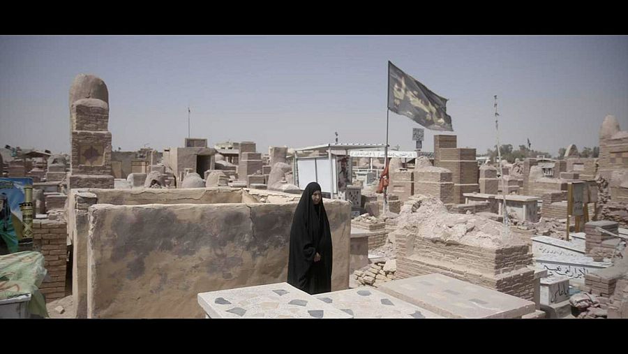 En un cementerio en ruinas, una mujer vestida de negro y con velo se encuentra entre tumbas de piedra dañadas, con una bandera oscura al fondo, transmitiendo una sensación de desolación.