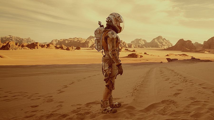 En un desierto de arena rojiza, un astronauta con un traje espacial blanco y naranja, y una mochila con el número 