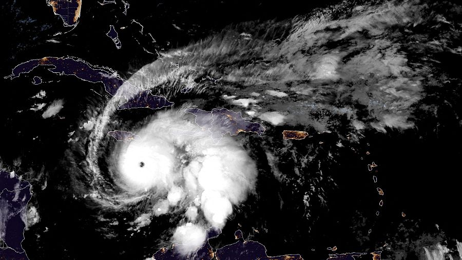 Vista satelital del huracán, una gran espiral de nubes blancas con un ojo oscuro en el centro, sobre el mar Caribe. Se observa una isla al oeste, iluminada.