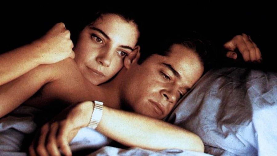 Una escena íntima muestra a Maribel Verdú y Jorge Sanz, jóvenes, en una cama. Maribel abraza a Jorge, quien tiene los ojos cerrados, mientras ella mira hacia arriba con expresión pensativa, en un ambiente de luz tenue.