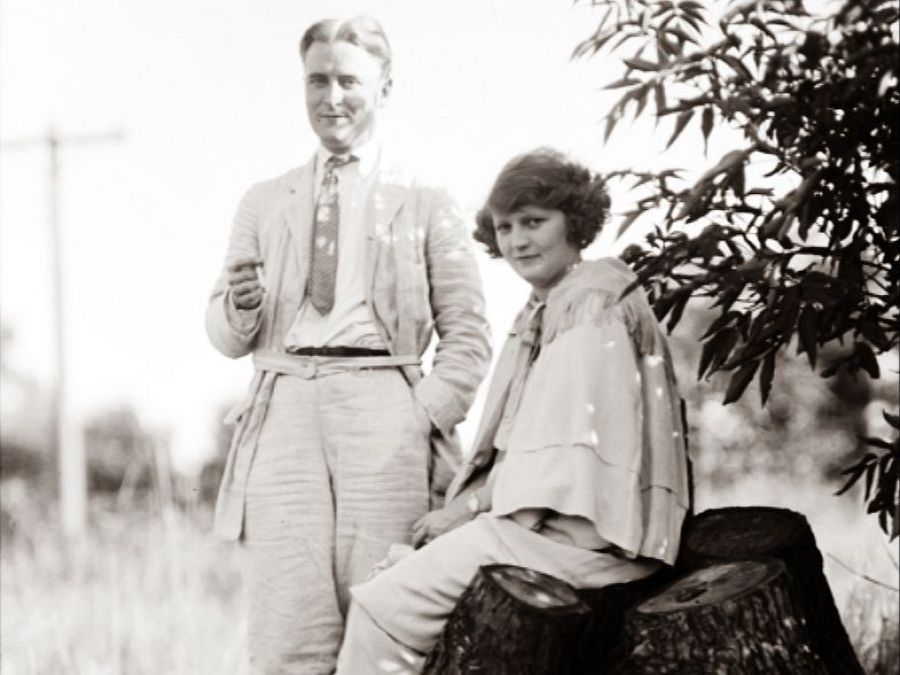 Una imagen en blanco y negro captura a un hombre con traje claro y una mujer con abrigo, posando en un entorno natural con un tronco de árbol. El hombre sostiene algo pequeño en la mano.