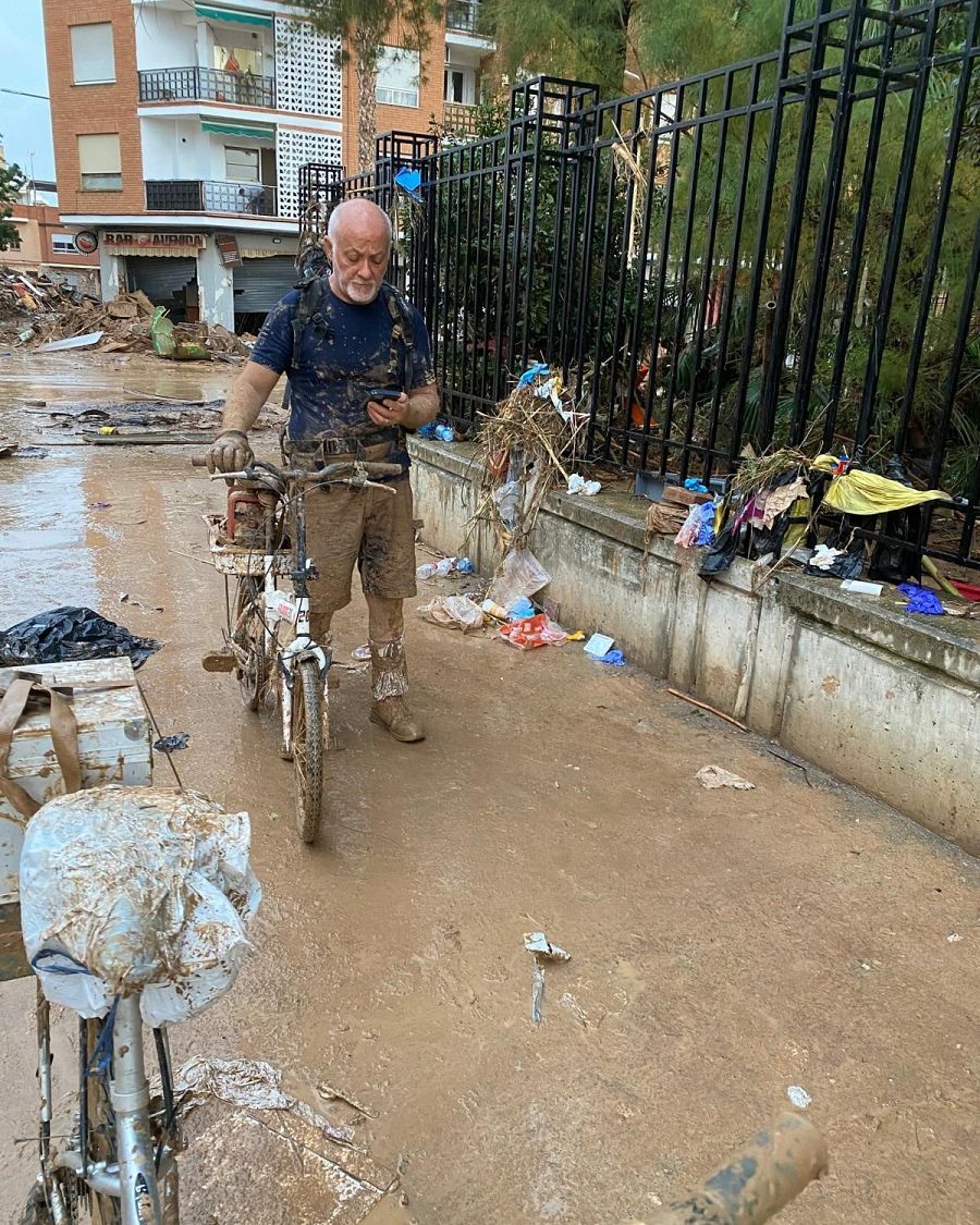 Un hombre con barba y calvicie, vestido con ropa oscura y botas, examina su teléfono móvil en una calle llena de barro, junto a una bicicleta también embarrada. Se observa un edificio al fondo y una valla metálica a la derecha.