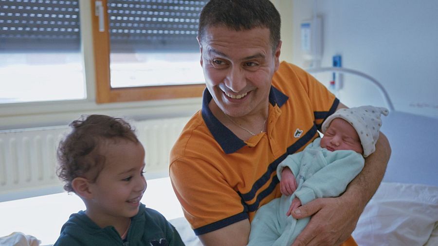 Un padre sostiene a su bebé recién nacido, mientras un niño pequeño observa la escena. El bebé está envuelto en una manta y lleva un gorro. La escena parece tener lugar en una habitación.