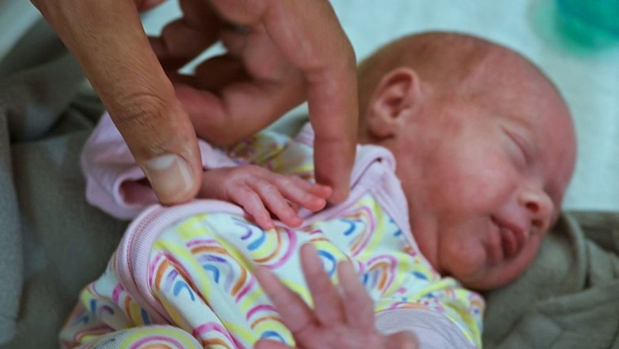 Un bebé recién nacido, envuelto en una manta, duerme plácidamente. Lleva un pijama blanco con un estampado de arcoíris y detalles rosas, mientras una mano adulta lo toca suavemente.