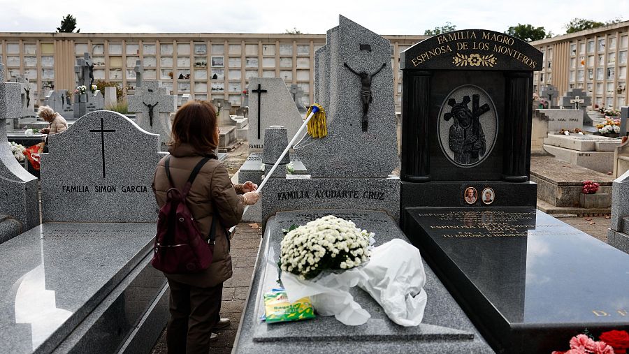 Una mujer se afana en la limpieza de una lápida con una fregona antes de colocar las flores.