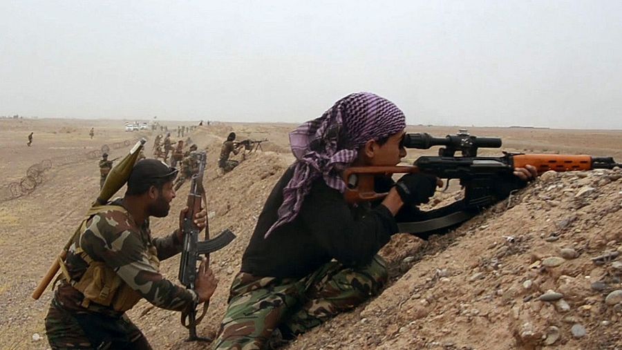 Un grupo de hombres armados se encuentra en un paisaje árido. Un hombre con pañuelo y guantes apunta con un rifle con mira telescópica, mientras que otro lleva un lanzacohetes y un rifle.