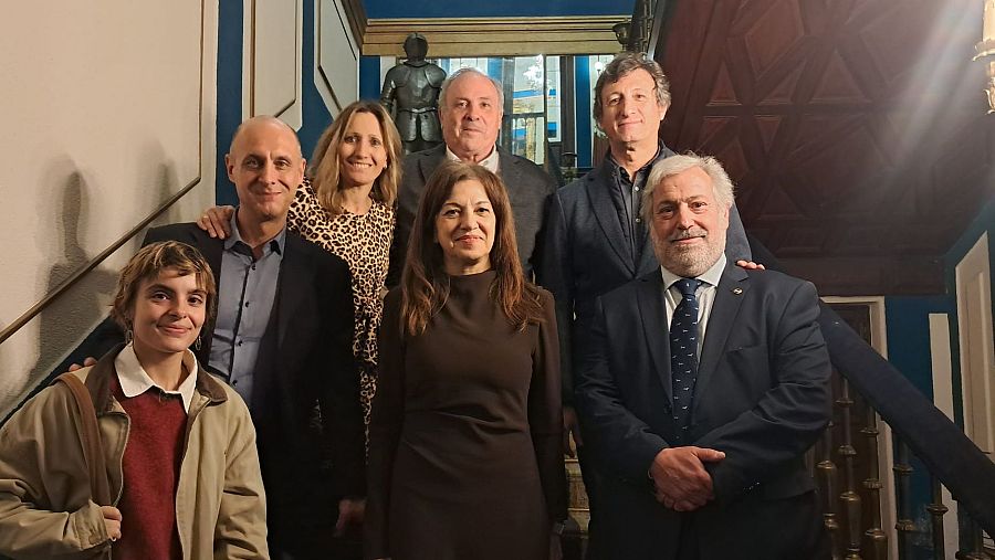 Un grupo de ocho personas, divididas en dos filas, posan en una escalera. Se observan diversas prendas de vestir y peinados, con una pared decorada al fondo.