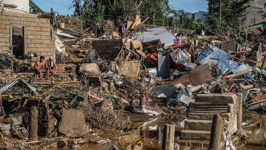 Una escena de desolación en una zona afectada por un tifón muestra escombros, edificios dañados y personas en medio del caos. El ambiente general refleja el impacto de una catástrofe natural.