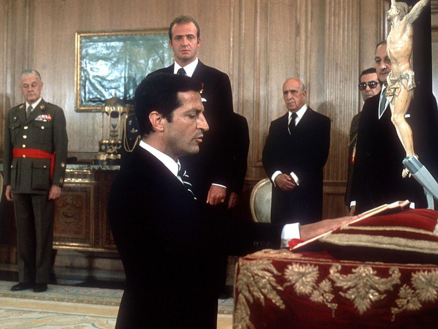 Adolfo Suárez, con la mano sobre una mesa cubierta por un paño, realiza el juramento presidencial. A su lado, el rey observa la ceremonia. Se distinguen otras personas vestidas formalmente y un militar con uniforme.