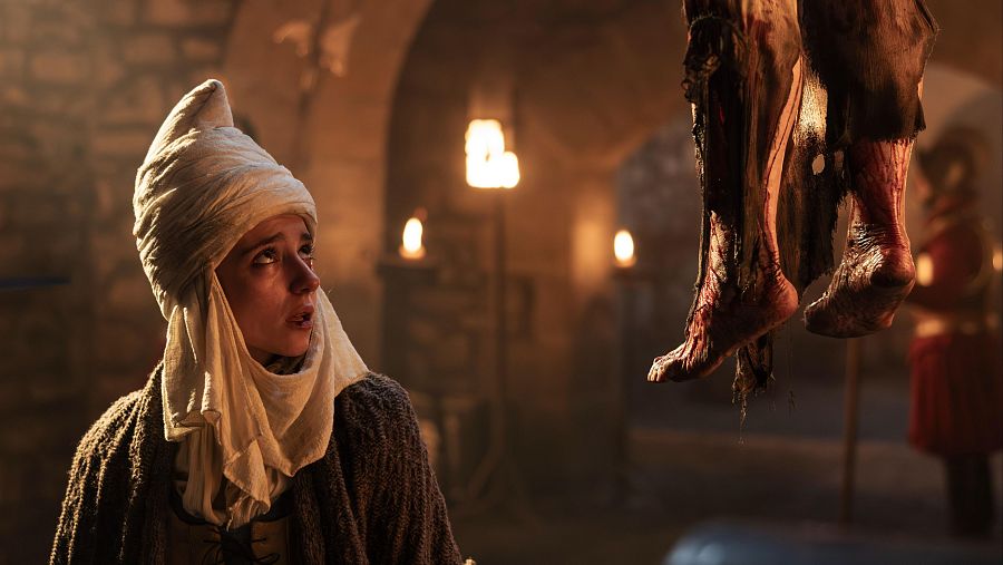 Una escena oscura muestra a una persona joven con turbante, posiblemente asustada, en un espacio iluminado por velas. En el fondo, se intuye la presencia de otra figura y, colgando, unos pies ensangrentados.