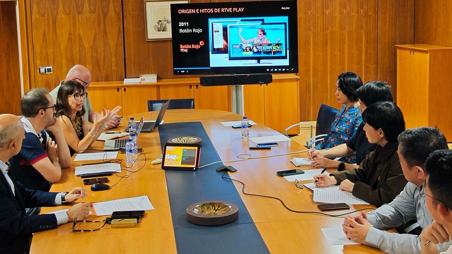 Una reunión entre dos delegaciones se lleva a cabo en una sala con paredes de madera. En la mesa, se observan computadoras portátiles, tabletas y documentos, mientras que en la pared se proyecta una presentación.