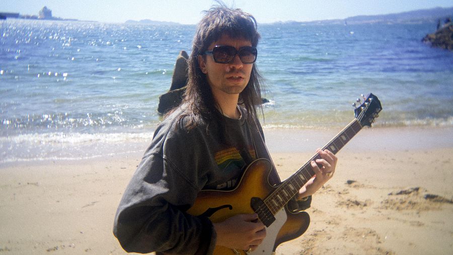 En una playa de arena clara, una persona con gafas de sol y sudadera gris toca una guitarra eléctrica, con el mar en calma y el cielo despejado como telón de fondo.