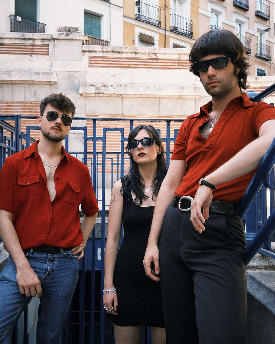 Tres individuos, incluyendo una mujer con vestido negro y dos hombres con camisas rojas, posan frente a una estructura con barandillas azules, posiblemente la entrada a un lugar subterráneo, con un edificio de piedra como telón de fondo.
