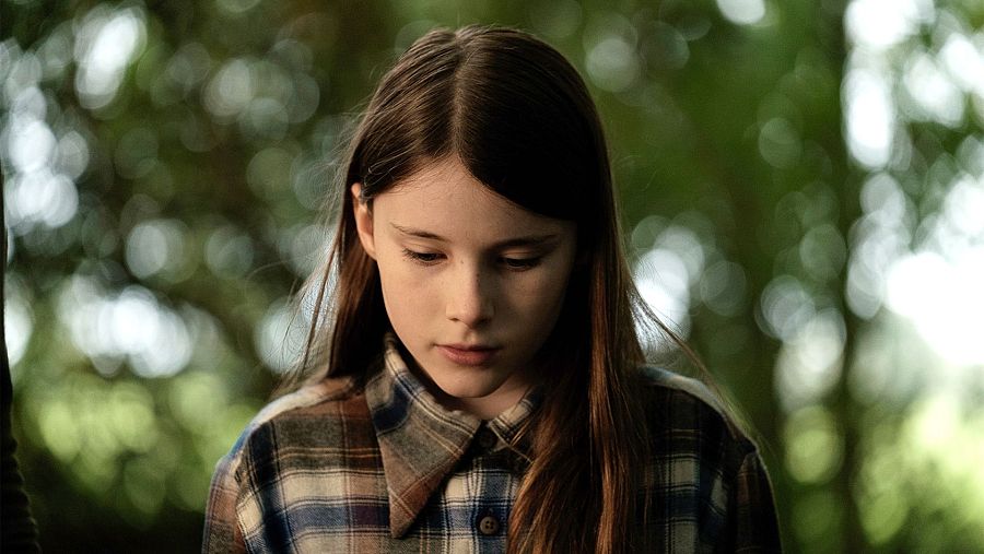 Una joven con cabello castaño y ojos claros, vestida con una camisa de cuadros, mira hacia abajo con una expresión melancólica en un entorno natural con vegetación borrosa.