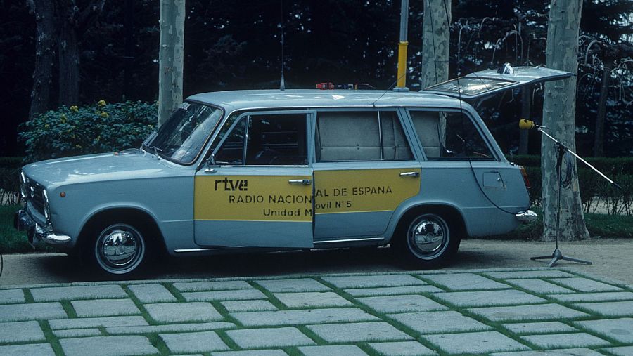 Un coche familiar azul claro, identificado como unidad móvil de Radio Nacional de España, con el maletero abierto y un micrófono preparado para su uso.