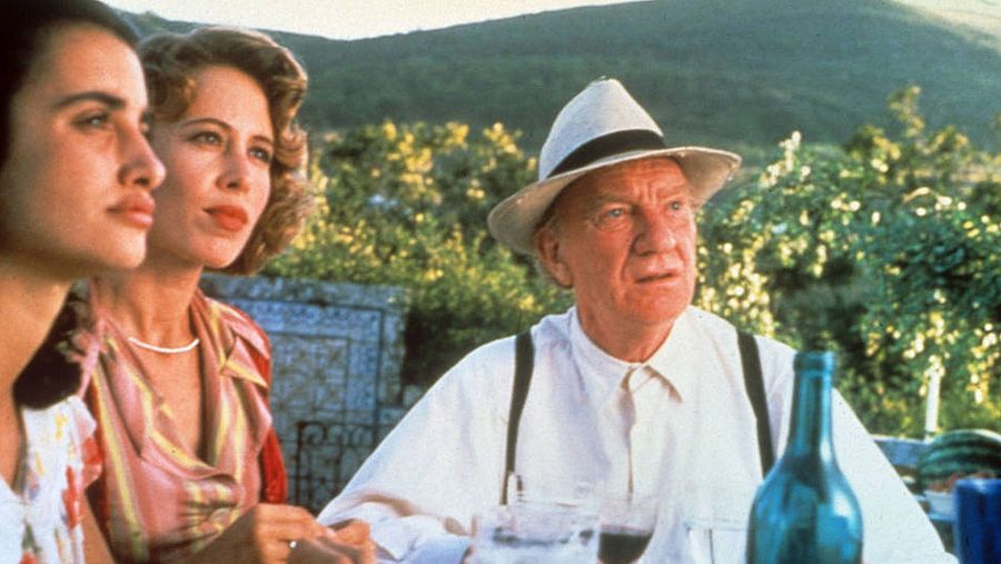 Tres personas, incluyendo una mujer de pelo oscuro y otra de pelo castaño claro, comparten una mesa con una botella de vino y una sandía en un entorno campestre. Un hombre mayor con sombrero y tirantes está presente.