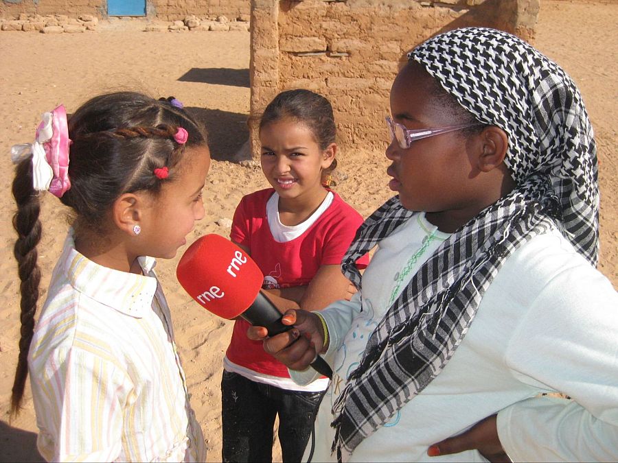 En un campamento desértico, dos niñas saharauis participan en una entrevista. Una de ellas, con una trenza y camisa a rayas, es entrevistada mientras otra sostiene un micrófono con el logo de 