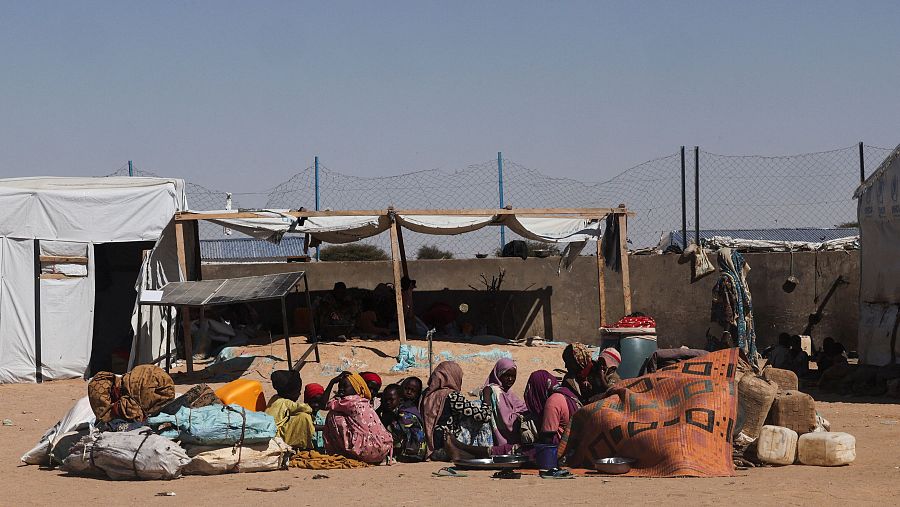Un grupo de personas, incluyendo mujeres y niños, se encuentran en un campamento improvisado en un entorno desértico, rodeados de sus pertenencias y con tiendas de campaña al fondo.