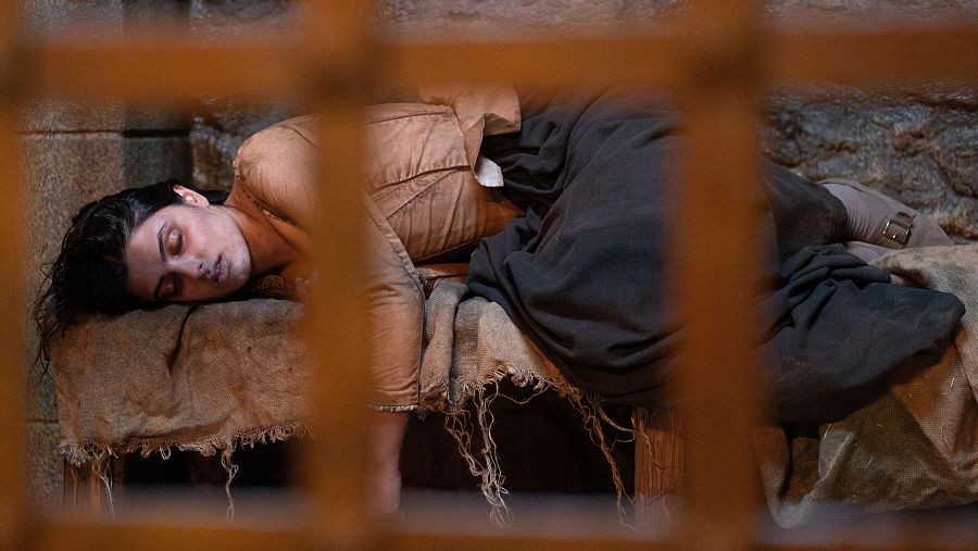 Una mujer llamada Luisa, con vestimenta oscura y cabello oscuro, yace inconsciente sobre una superficie cubierta con telas rústicas, en un entorno que parece ser una celda o lugar similar, con una reja visible.