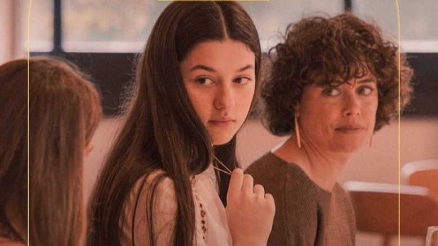 En la imagen se observan dos mujeres en un interior. Una de ellas lleva un jersey marrón y pendientes largos, mientras que la otra viste una blusa blanca con detalles bordados y un collar.