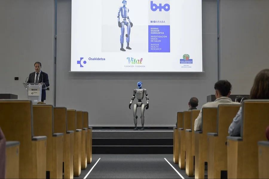 Un robot humanoide gris y azul, llamado SAAKI, se presenta en una plataforma durante un evento. Un hombre, posiblemente un consejero de salud, habla desde un atril frente a una audiencia, con una pantalla que muestra logos y texto sobre investigación.
