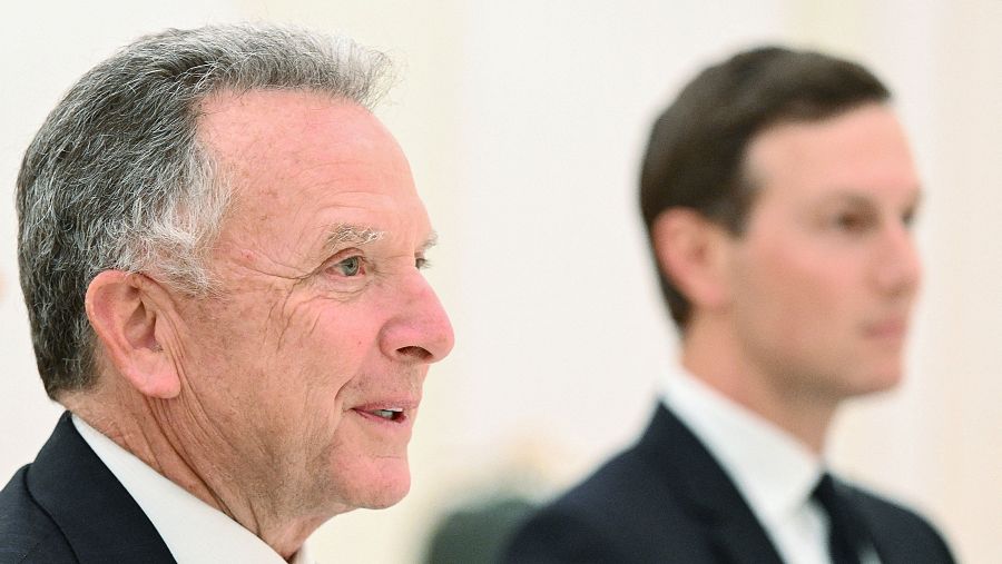 En la imagen, dos hombres de perfil, ambos con trajes oscuros, se encuentran uno frente al otro. Uno de ellos, de mayor edad, tiene cabello gris y piel clara.