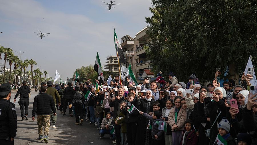 En una calle de Damasco, una multitud celebra con banderas y teléfonos móviles. Se observan soldados y civiles en un ambiente festivo, con helicópteros en el cielo.