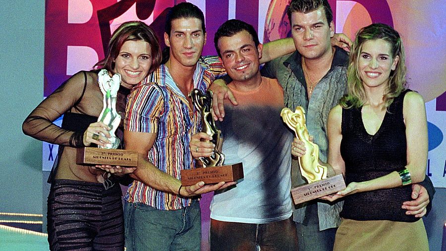 Cinco personas posan con trofeos; una mujer con blusa negra y pantalones de cebra, un hombre con camisa de rayas, otro con camiseta degradada, un hombre con camisa gris y una mujer rubia con falda café. El fondo es difuso con colores morados y rosados.