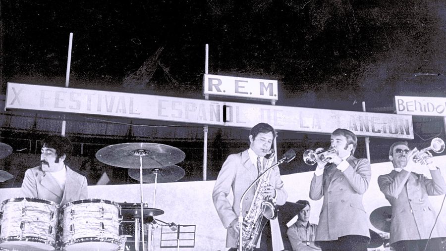 En un escenario, cuatro músicos vestidos con trajes actúan durante un festival. Se observa a un baterista, un saxofonista y dos trompetistas tocando sus instrumentos, con un letrero que anuncia el evento.