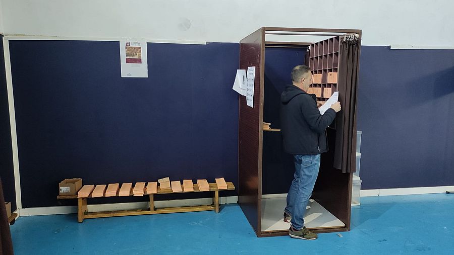 Un hombre con chaqueta oscura y zapatillas deportivas revisa un papel dentro de una cabina de votación de madera con cortinas. En el exterior, se aprecian papeletas y una caja, en un espacio interior con paredes claras.