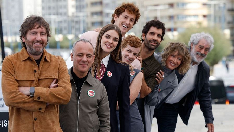 Ocho individuos sonríen a la cámara en una fotografía al aire libre. Se distinguen por su vestimenta diversa, incluyendo chaquetas y uniformes, con un fondo de edificios y un cielo nublado.